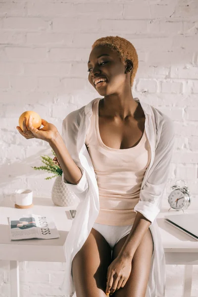 Cheerful african american woman with short hair holding orange in kitchen — Stock Photo