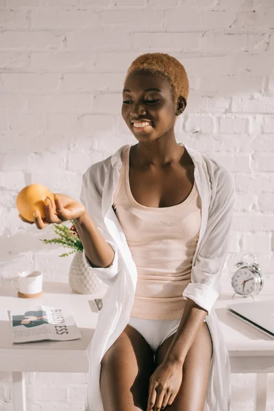 Mujer afro-americana feliz con pelo corto sosteniendo naranja en la cocina - foto de stock