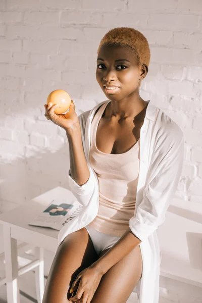 Attraktive afrikanisch-amerikanische Frau mit kurzen Haaren hält orange in weißer Küche — Stockfoto