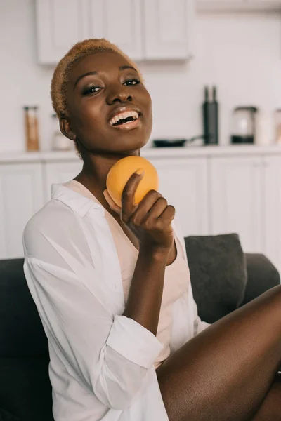 Mujer afro-americana feliz con pelo corto sosteniendo naranja en la sala de estar - foto de stock