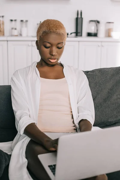 Mujer afroamericana concentrada escribiendo en el ordenador portátil en la sala de estar - foto de stock