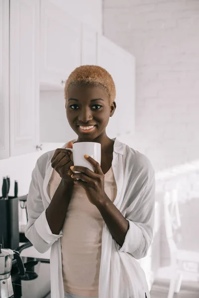 Alegre mujer afroamericana con taza de café en cocina blanca - foto de stock