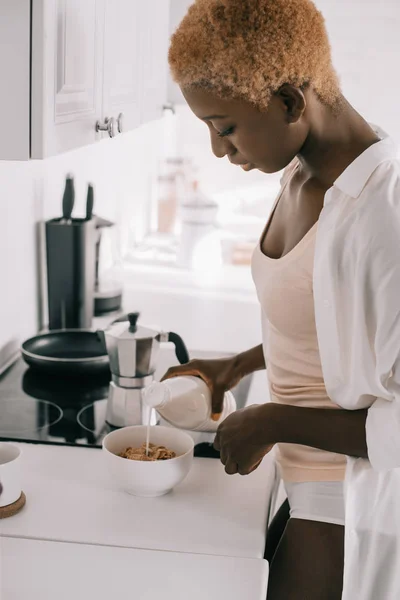 Bella donna afroamericana versando latte in ciotola con cornflakes in cucina — Foto stock