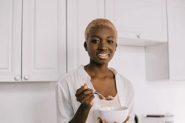 Mujer afroamericana feliz sosteniendo cuchara y tazón con copos de maíz - foto de stock