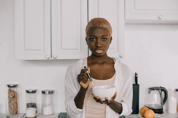 Confiante Africano americano mulher segurando colher e tigela com flocos de milho na cozinha branca — Fotografia de Stock