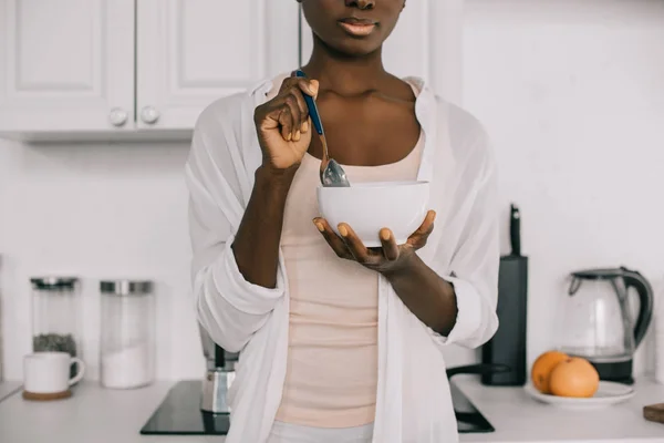 Vista cortada de mulher afro-americana segurando colher e tigela com flocos de milho na cozinha branca — Fotografia de Stock