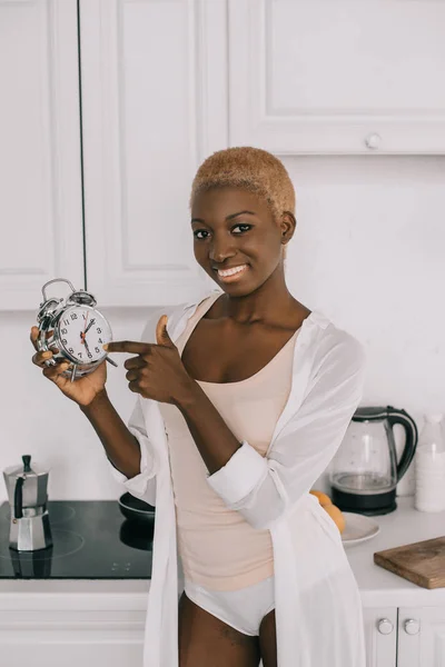 Веселая африканская американка, указывающая на часы на белой кухне — стоковое фото