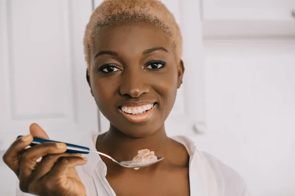 Nahaufnahme einer fröhlichen afrikanisch-amerikanischen Frau, die einen Löffel mit Cornflakes in der weißen Küche hält — Stockfoto