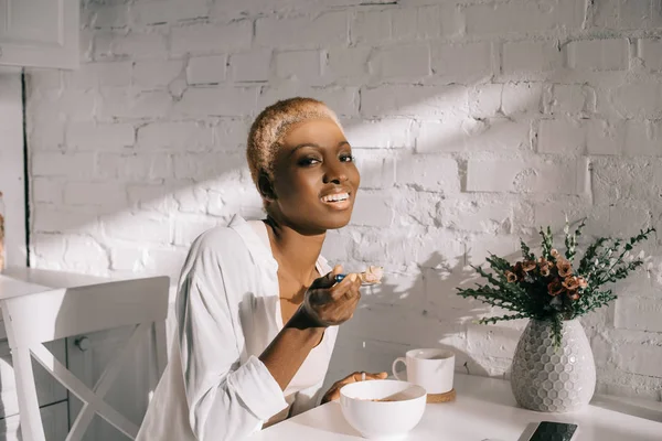 Mulher americana africana alegre segurando colher com flocos de milho e sorrindo na cozinha — Fotografia de Stock