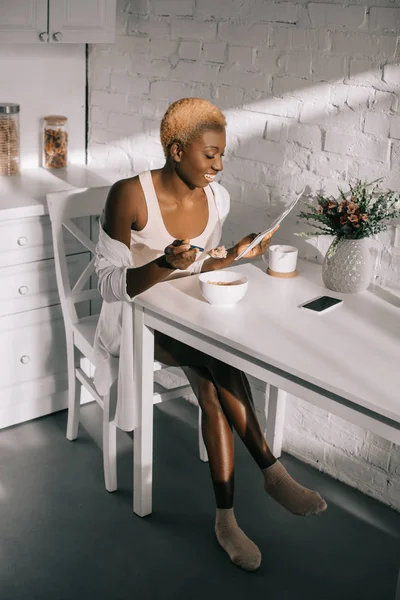 Cheerful african american woman reading newspaper and having breakfast in white kitchen — Stock Photo