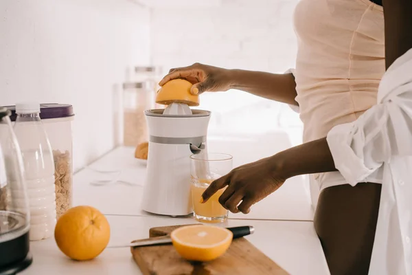 Vista recortada de mujer afroamericana preparando jugo de naranja fresco en cocina blanca - foto de stock