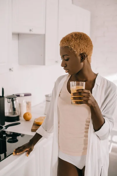 Thoughtful african american woman holding glass of orange juice in kitchen — Stock Photo