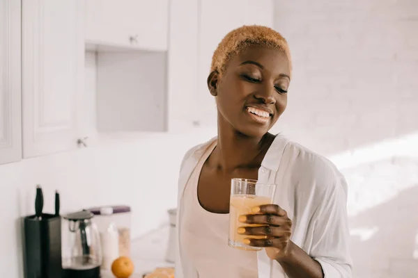 Cheerful african american woman holding glass of orange juice in kitchen — Stock Photo