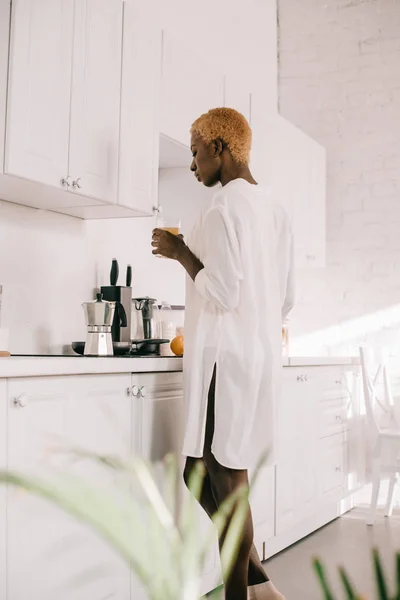 Mulher americana africana com cabelo curto segurando vidro de suco de laranja na cozinha branca — Fotografia de Stock