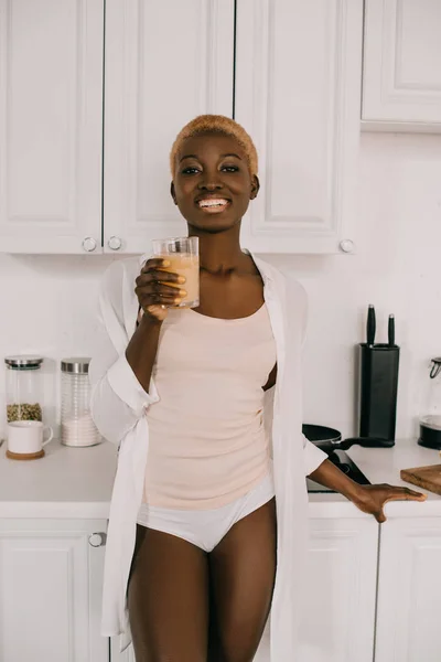 Heureuse femme afro-américaine montrant verre de jus d'orange dans la cuisine blanche — Photo de stock