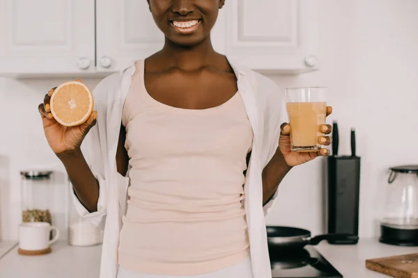 Vista recortada de mujer afroamericana mostrando vaso de jugo de naranja y la mitad de naranja - foto de stock