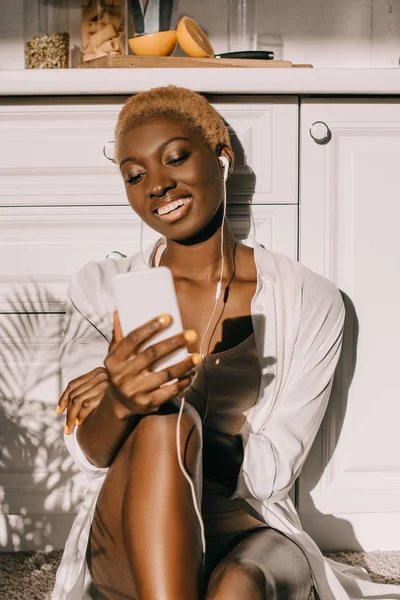 Femme afro-américaine gaie tenant smartphone et écouter de la musique dans les écouteurs dans la cuisine blanche — Photo de stock