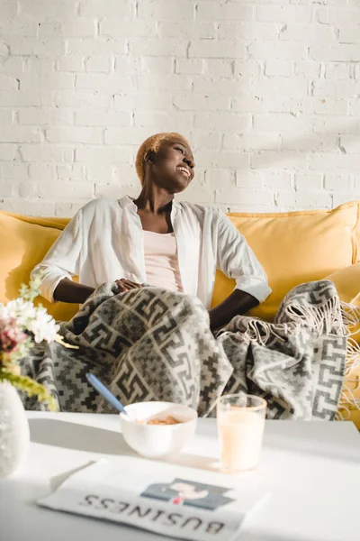 Mujer afroamericana feliz descansando debajo de una manta en un sofá amarillo - foto de stock