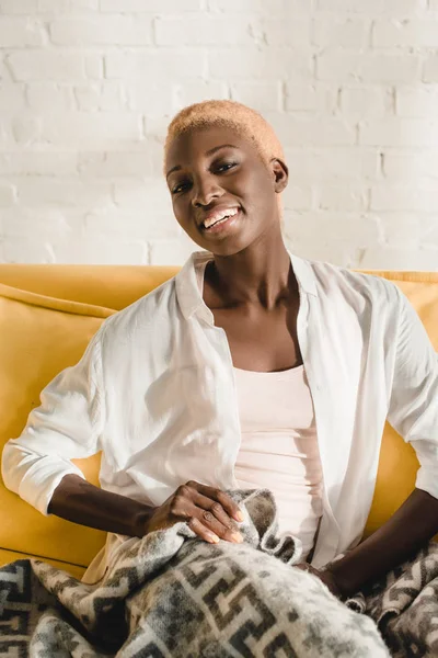 Cheerful african american woman relaxing under blanket on yellow sofa — Stock Photo