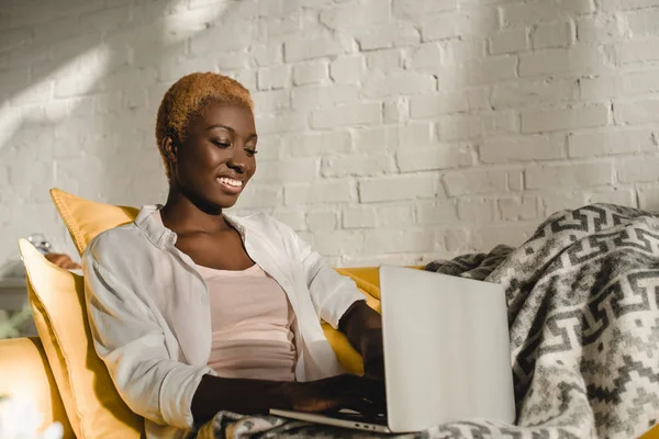 Cheerful african american woman using laptop on yellow sofa — Stock Photo