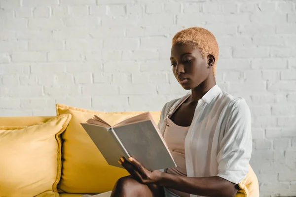 Attractive african american woman with short hair reading book on yellow sofa — Stock Photo