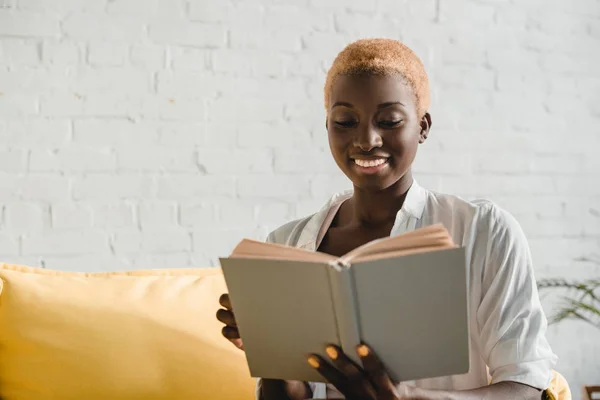 Mulher americana africana alegre com cabelo curto leitura livro no sofá amarelo — Fotografia de Stock