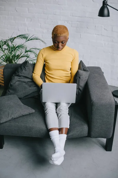 Femme afro-américaine concentrée avec les cheveux courts assis sur le canapé et en utilisant un ordinateur portable — Photo de stock