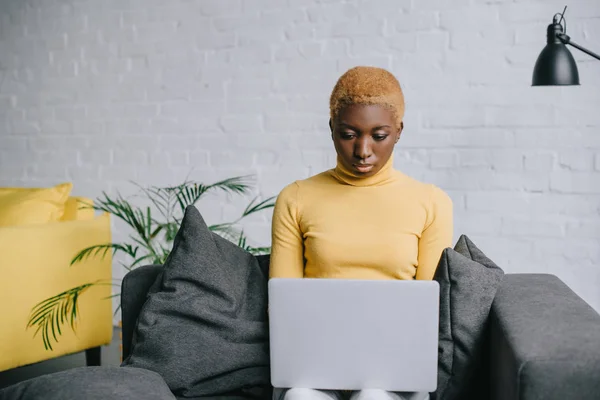 Konzentrierte Afroamerikanerin benutzt Laptop während sie auf dem Sofa sitzt — Stockfoto