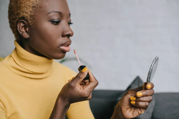 Pensive african american woman applying lip gloss and looking in mirror — Stock Photo