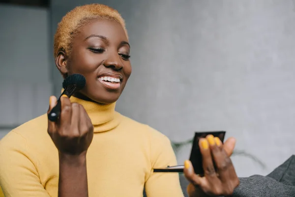 Heureuse femme afro-américaine appliquant la poudre avec pinceau cosmétique — Photo de stock