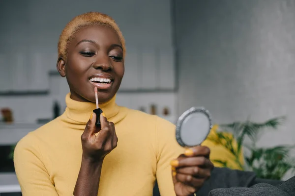 Веселая африканская американка, надевающая блеск для губ и смотрящая в зеркало — стоковое фото