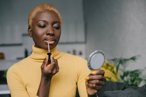 Concentrated african american woman applying lip gloss and looking in mirror — Stock Photo