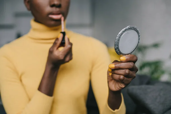 Selective focus of african american woman holding mirror and applying lip gloss — Stock Photo