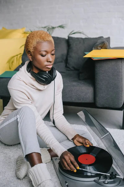 Mujer afroamericana soñadora sentada en la alfombra en auriculares cerca del reproductor de discos - foto de stock