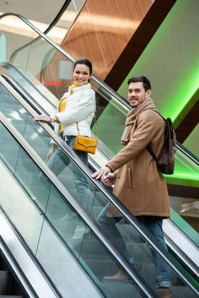 Handsome man and beautiful girl smiling and going up on escalator in shopping mall — Stock Photo