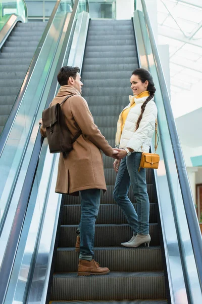 Coppia sorridente in abiti caldi che si tiene per mano, guardandosi l'un l'altro e salendo sulla scala mobile — Foto stock