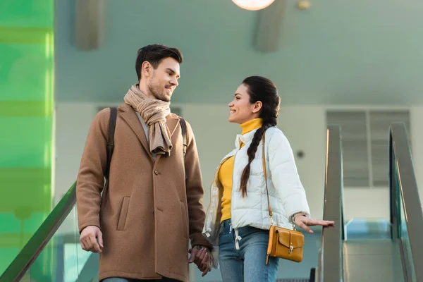 Attractive couple holding hands, looking at each other and going down on escalator — Stock Photo