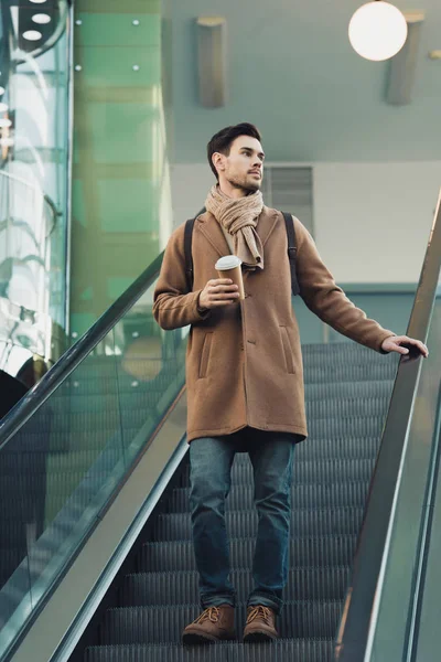 Handsome man going down on escalator and holding disposable cup — Stock Photo