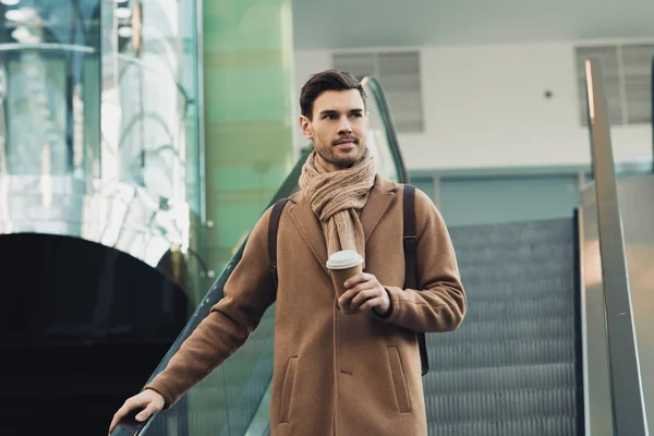 Schöner Mann in warmer Kleidung hält Pappbecher in der Hand und geht auf Rolltreppe hinunter — Stockfoto