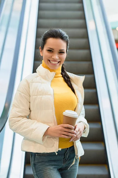 Hermosa chica bajando en escaleras mecánicas y sosteniendo taza de papel - foto de stock