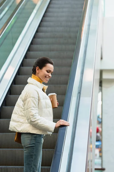 Hermosa chica sonriendo, subiendo en escaleras mecánicas y sosteniendo taza desechable - foto de stock