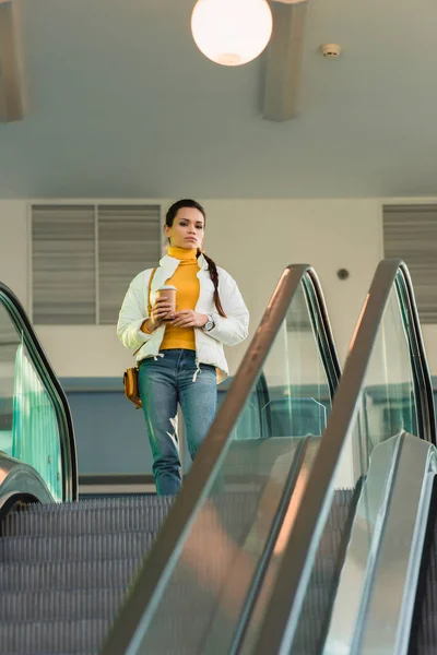 Chica atractiva con taza desechable de pie en la parte superior de la escalera mecánica - foto de stock