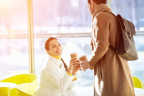 Приваблива сидяча дівчина в теплому одязі і стоячий чоловік тримає одноразові чашки в кафе — стокове фото