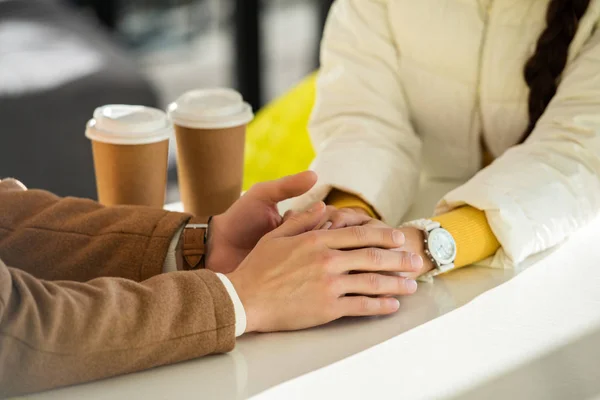 Cropped view of loving couple holding hands at table with two disposable cups — Stock Photo