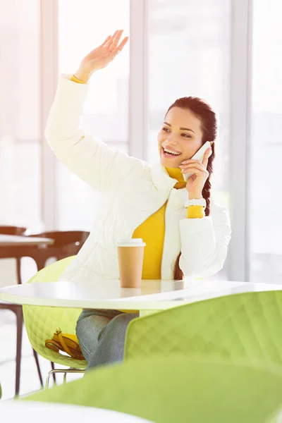 Smiling girl sitting at table, talking on smartphone and waving with hand in cafe — Stock Photo