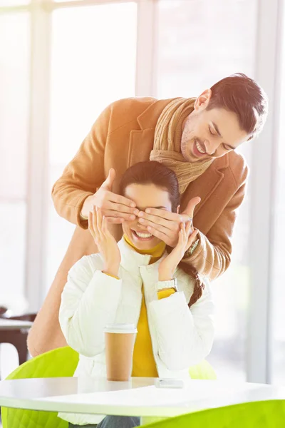 Schöner Mann schließt Augen lächelnder Freundin mit Händen, während Frau am Tisch im Café sitzt — Stockfoto