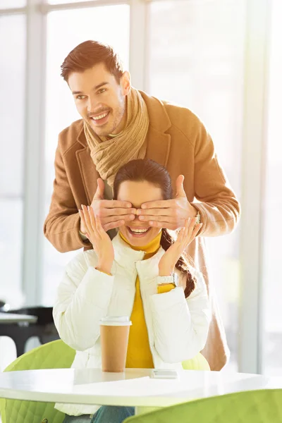 Homem bonito sorrindo e fechando os olhos da namorada feliz com as mãos, enquanto a mulher sentada à mesa no café — Fotografia de Stock