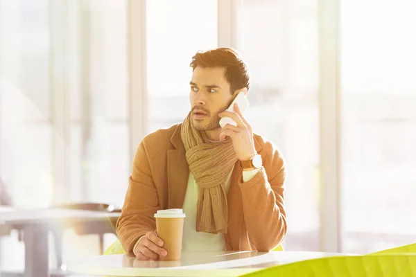 Besorgter Mann sitzt am Tisch, hält Pappbecher in der Hand und telefoniert im Café mit Smartphone — Stockfoto