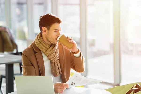 Bel homme d'affaires assis à table, buvant du café dans une tasse en papier et lisant un journal dans un café — Photo de stock