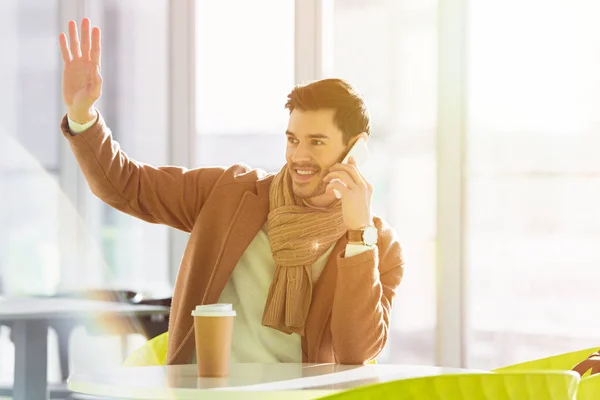 Hombre sonriente sentado en la mesa, hablando en el teléfono inteligente y saludando con la mano en el café - foto de stock